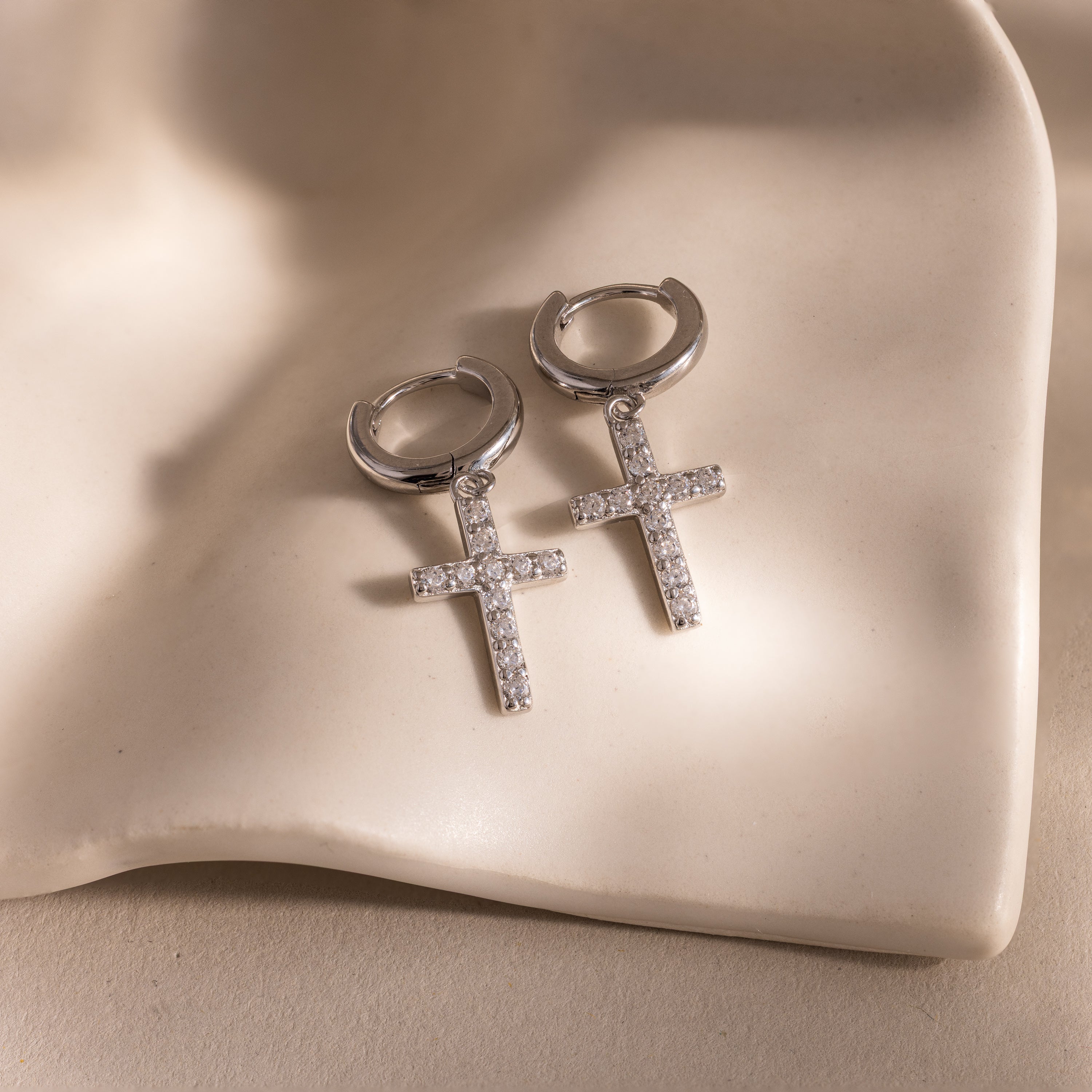 Flat-lay of silver huggie hoop earrings with dangling pave cross charms, displayed on a wavy ceramic surface.