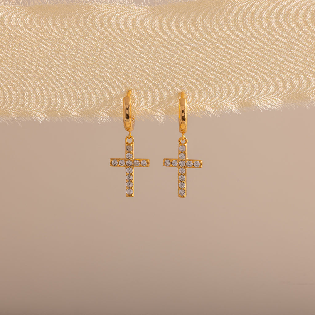 Gold huggie hoop earrings with dangling pave cross charms, hanging on a neutral fabric trim.