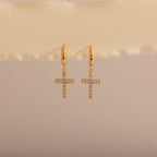 Gold huggie hoop earrings with dangling pave cross charms, hanging on a neutral fabric trim.