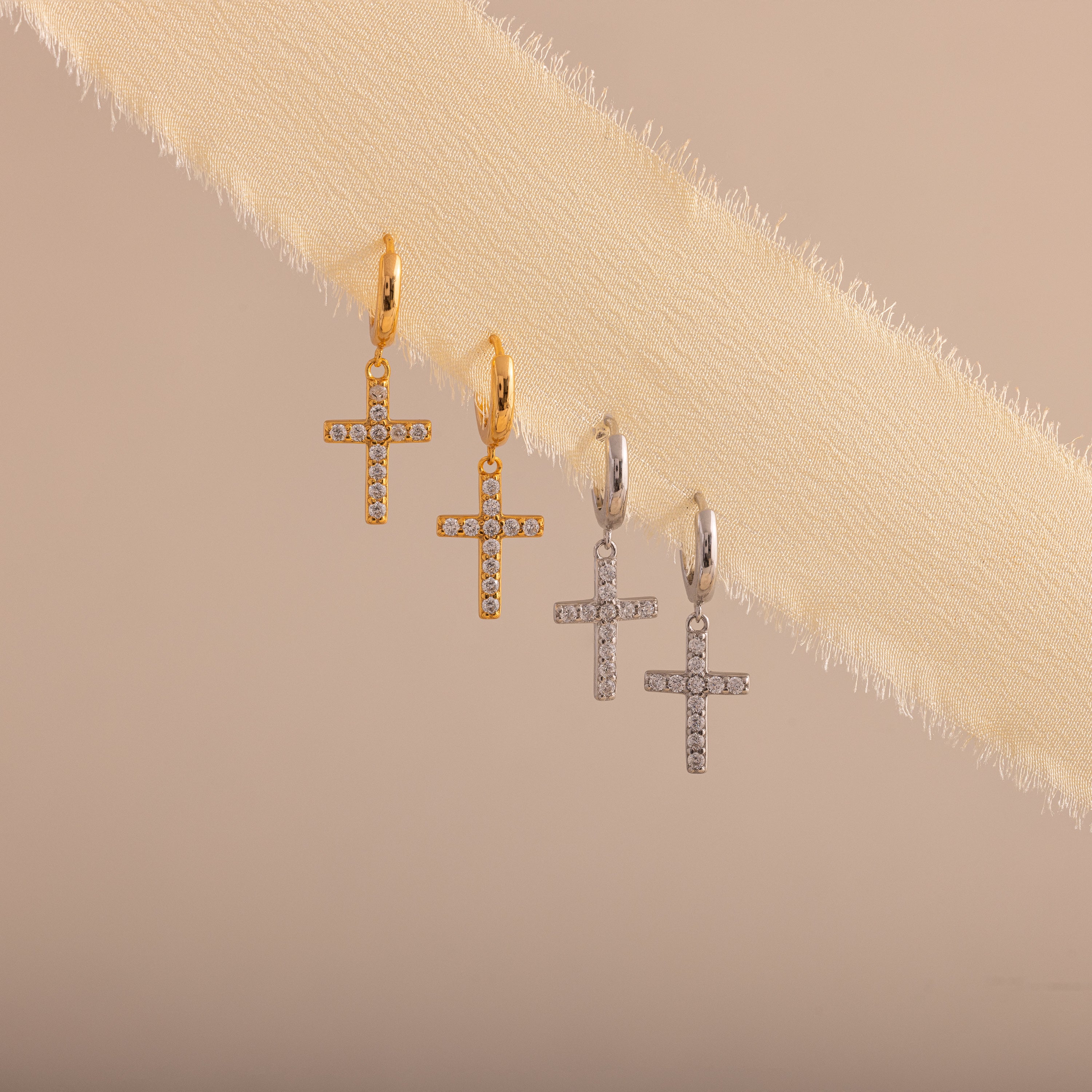 Gold and silver hoop earrings with dangling cross charms adorned with sparkling pave stones, displayed diagonally on a soft beige fabric background.