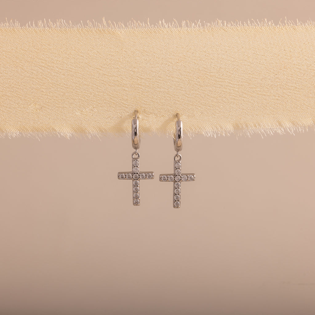 Silver huggie hoop earrings with dangling pave cross charms, hanging on a soft beige fabric.