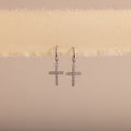 Silver huggie hoop earrings with dangling pave cross charms, hanging on a soft beige fabric.