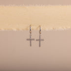 Silver huggie hoop earrings with dangling pave cross charms, hanging on a soft beige fabric.