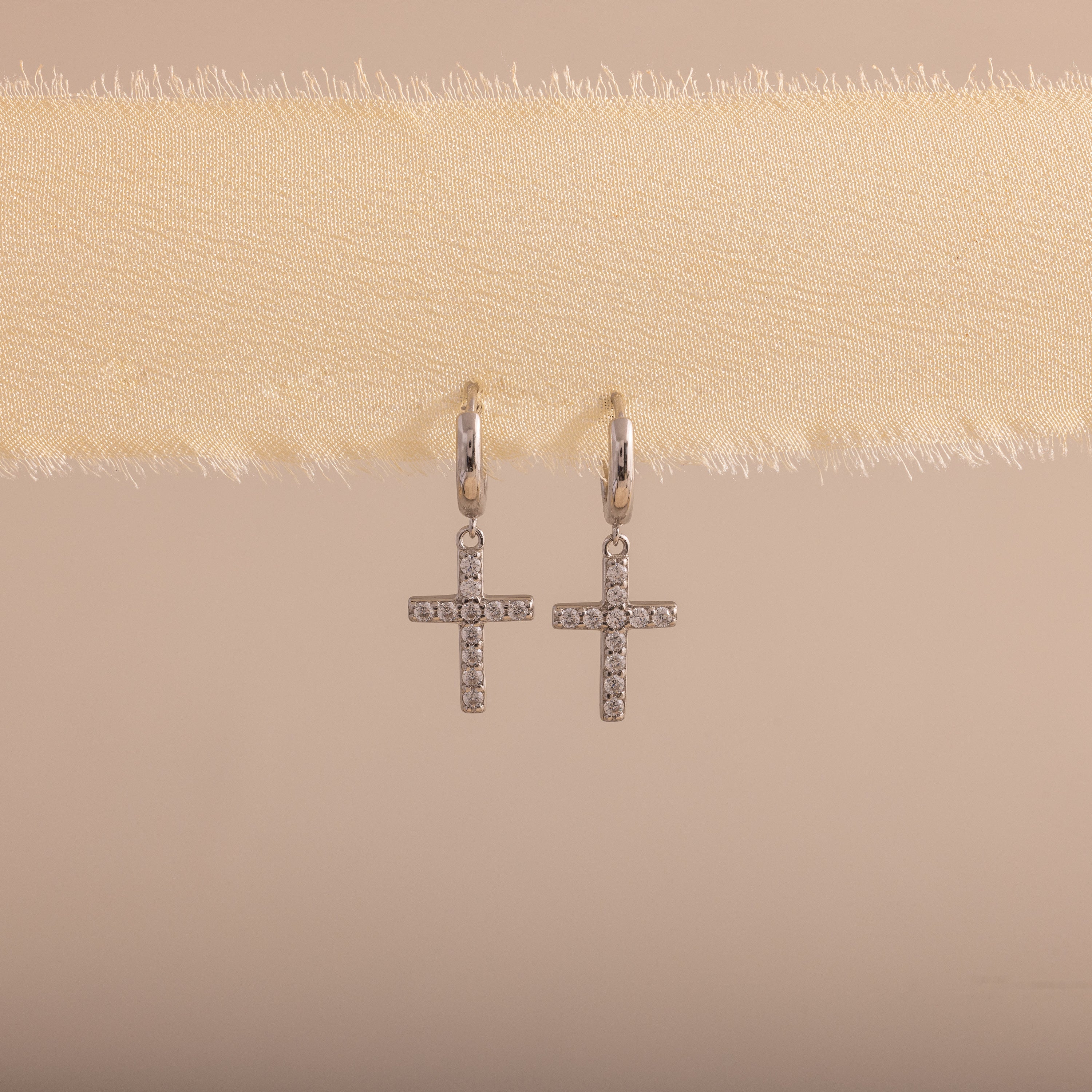 Silver huggie hoop earrings with dangling pave cross charms, hanging on a soft beige fabric.