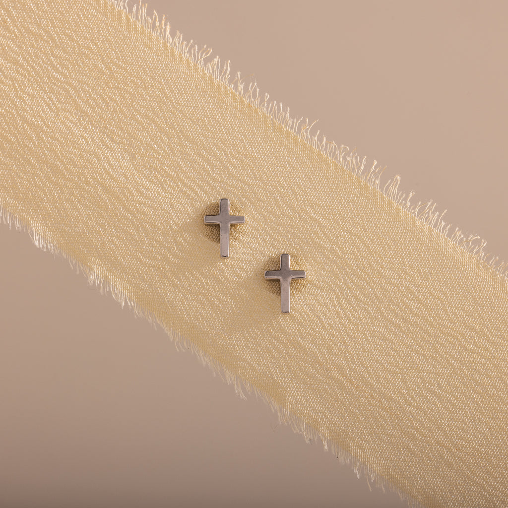 Dainty silver cross stud earrings displayed on a neutral ribbon fabric.