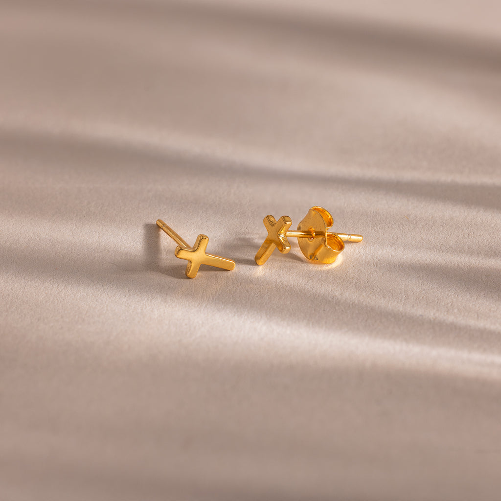 Pair of gold cross stud earrings with butterfly backings, displayed on a soft beige surface.