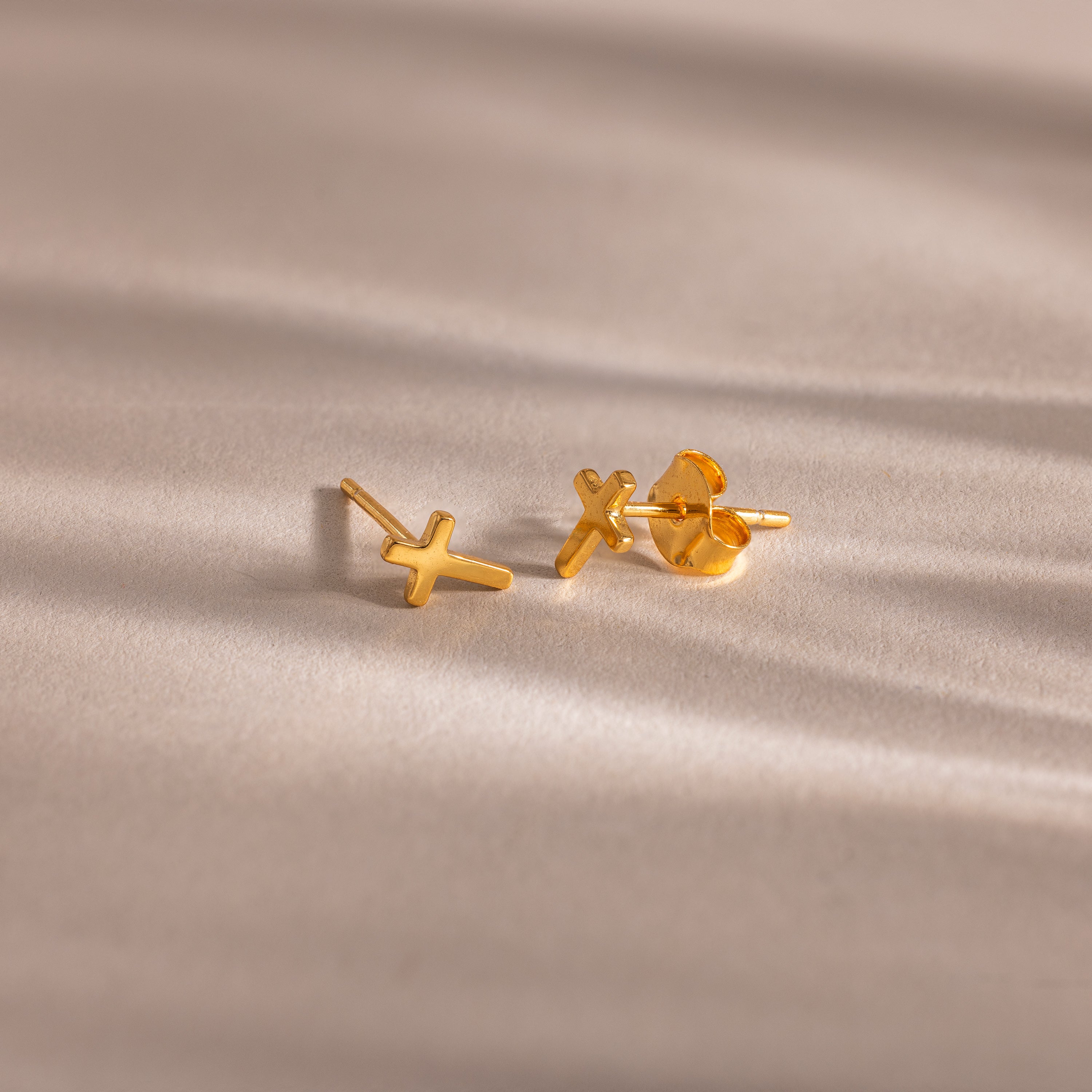 Pair of gold cross stud earrings with butterfly backings, displayed on a soft beige surface.