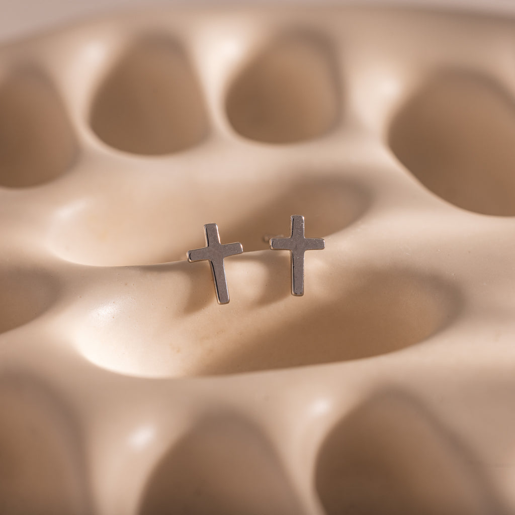 Close-up of dainty silver cross stud earrings, displayed on a neutral stone surface.