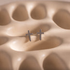 Close-up of dainty silver cross stud earrings, displayed on a neutral stone surface.