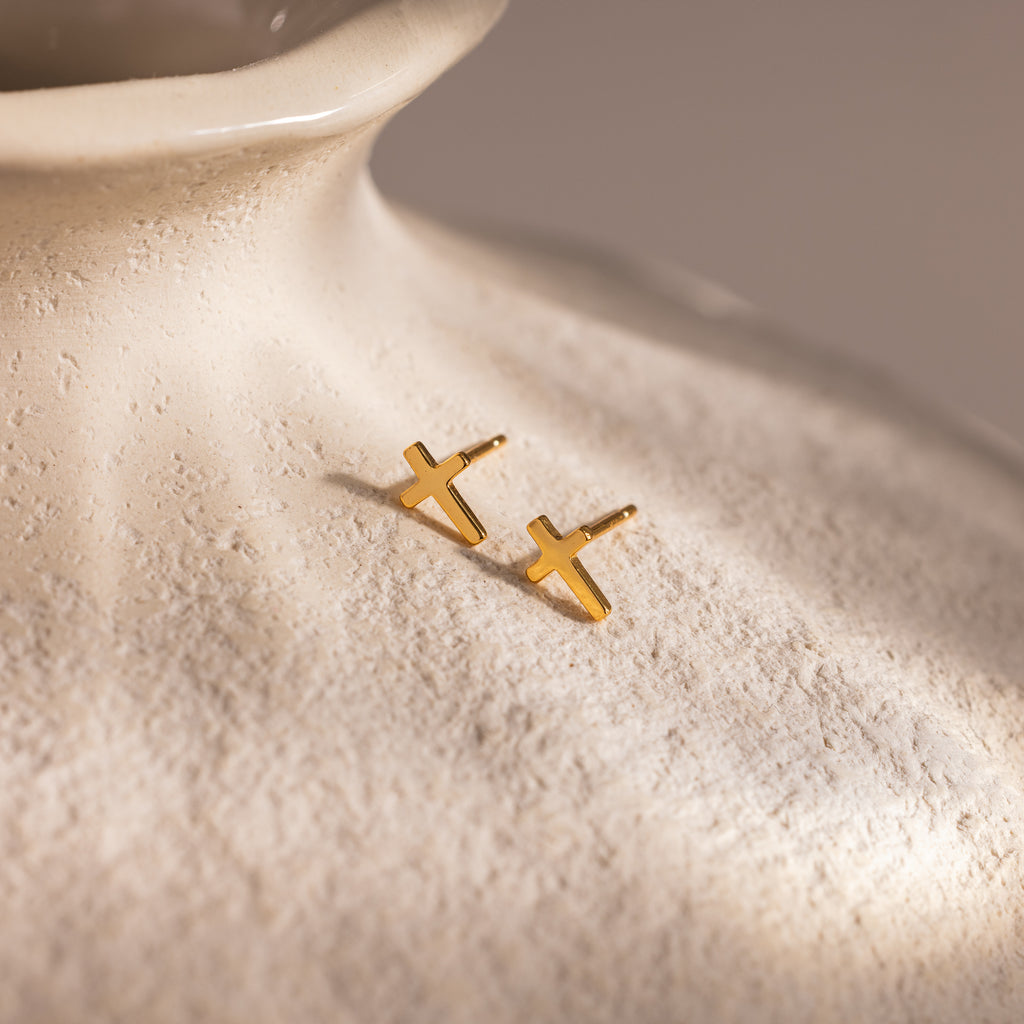 Pair of gold cross stud earrings displayed on the textured surface of a cream ceramic vase.
