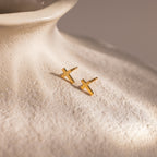 Pair of gold cross stud earrings displayed on the textured surface of a cream ceramic vase.