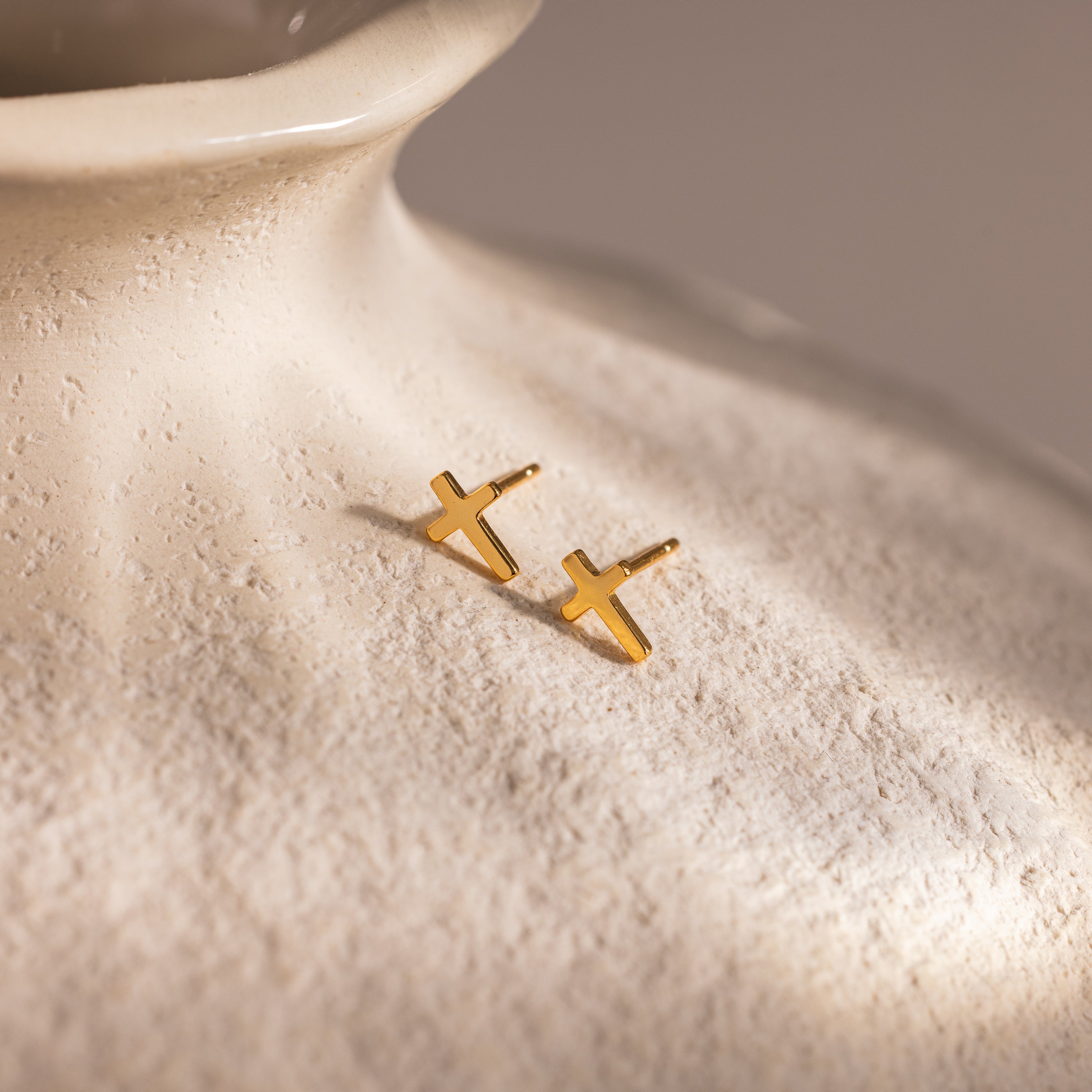Pair of gold cross stud earrings displayed on the textured surface of a cream ceramic vase.