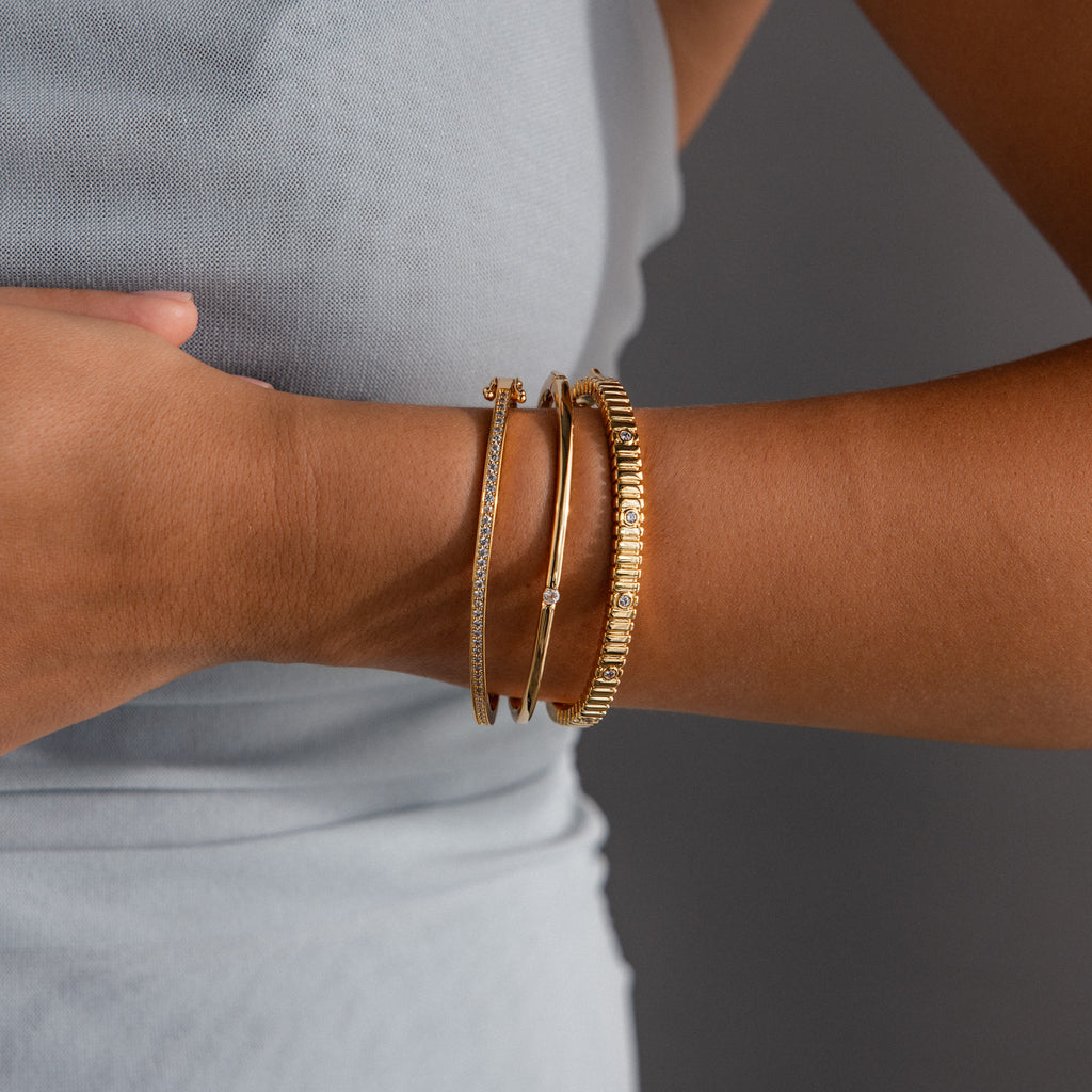 A person wearing three gold bracelets, including the Ribbed Diamond Bangle in 18K Gold, on their wrist against a light grey sleeveless top.