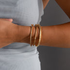 A person wearing three gold bracelets, including the Ribbed Diamond Bangle in 18K Gold, on their wrist against a light grey sleeveless top.