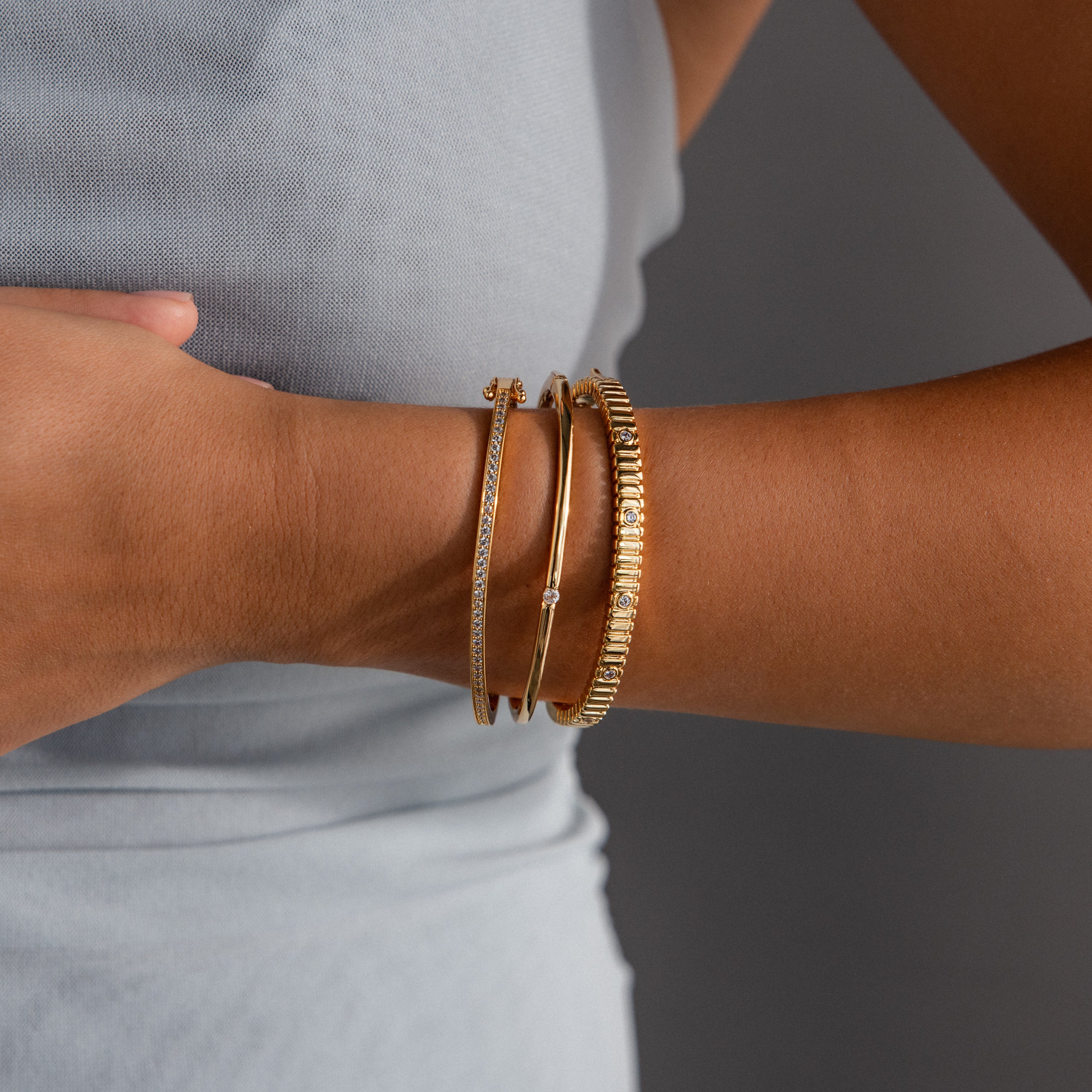 A person wearing three gold bracelets, including the Ribbed Diamond Bangle in 18K Gold, on their wrist against a light grey sleeveless top.