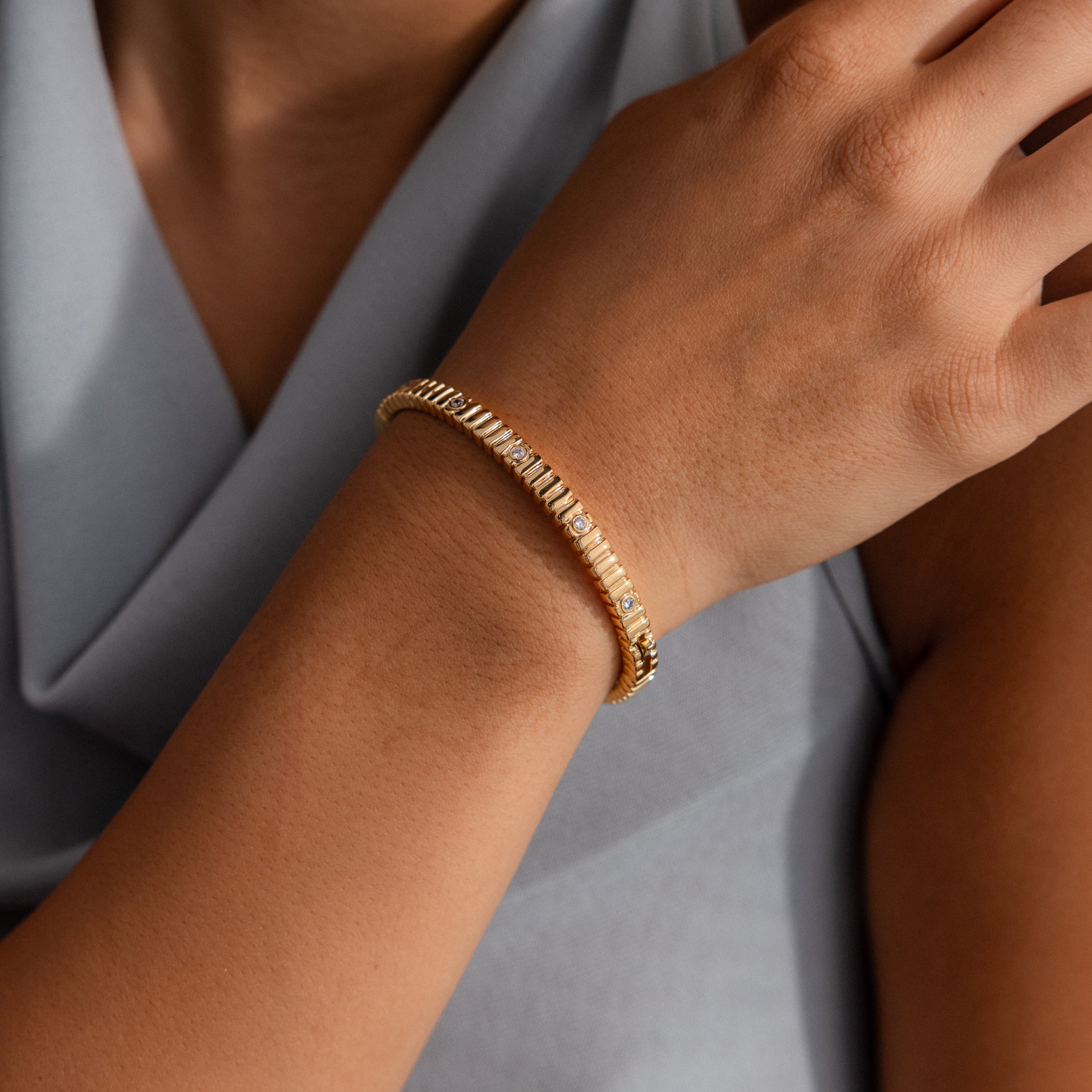 The Ribbed Diamond Bangle, featuring a polished gold finish and bezel set diamonds, adds elegance to a wrist when styled with a light gray sleeveless top.