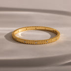 The Ribbed Diamond Bangle in 18K Gold features grooved edges and sparkling diamonds, elegantly displayed on a soft beige surface.