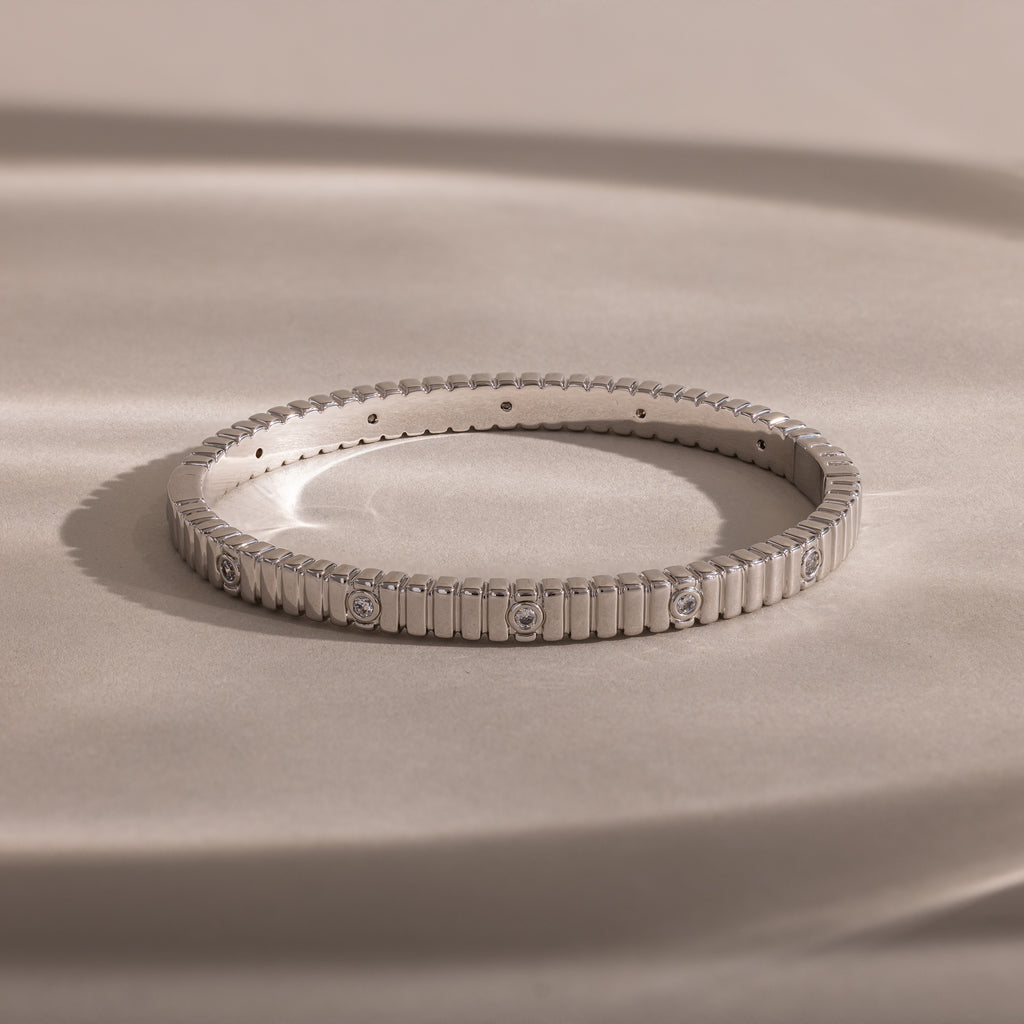 The Ribbed Diamond Bangle in White Gold, featuring small bezel set diamonds, rests on a smooth beige surface under soft lighting.