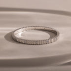 The Ribbed Diamond Bangle in White Gold, featuring small bezel set diamonds, rests on a smooth beige surface under soft lighting.