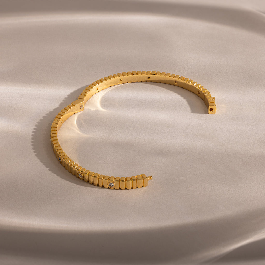 The Ribbed Diamond Bangle in 18K Gold features a ridged design and sparkling diamonds, elegantly arranged in a semi-circle on a soft beige surface.