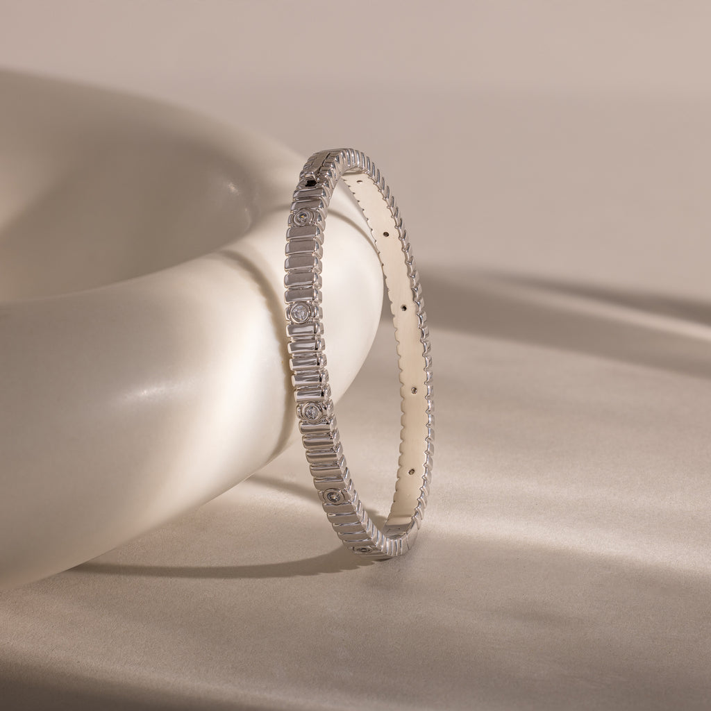 The Ribbed Diamond Bangle in White Gold, featuring a polished finish and rectangular details, rests against a smooth, curved white surface on a beige background.