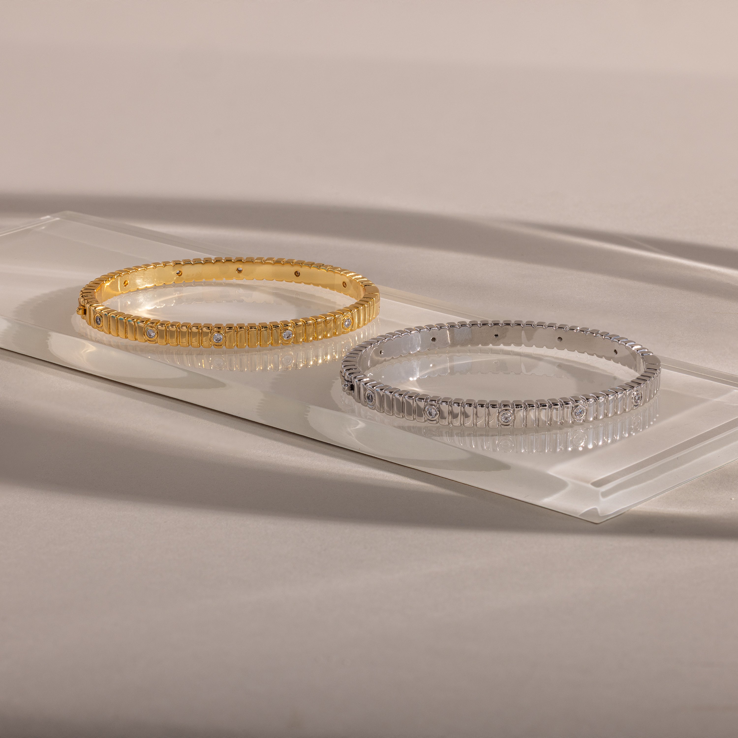 A gold and a silver Ribbed Diamond Bangle with a polished finish are displayed on a clear rectangular glass platform, one showcasing an elegant ribbed design for extra sparkle.