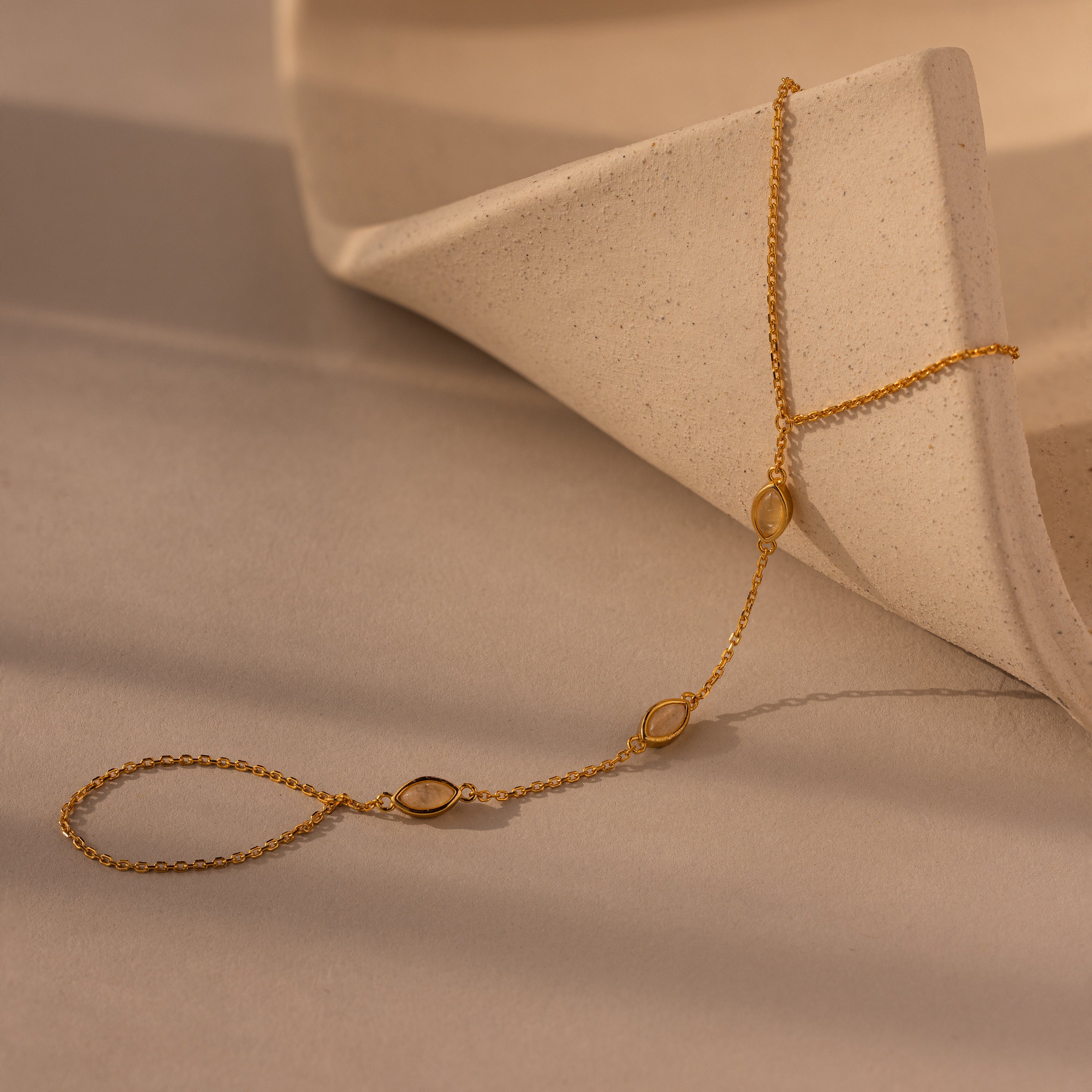 The Moonstone Hand Chain Bracelet in 18K Gold features delicate oval accents and is displayed on a curved, textured beige surface—an elegant celestial-inspired jewelry piece.