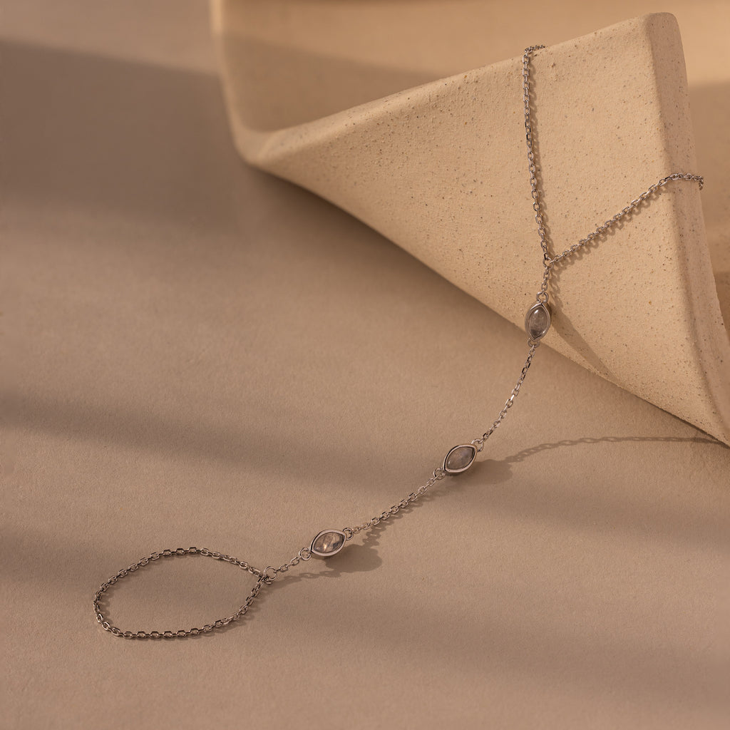 The Moonstone Hand Chain Bracelet in Silver features three small moonstones on a delicate chain, beautifully displayed on a curved, neutral-toned ceramic surface—an elegant piece of celestial-inspired jewelry.