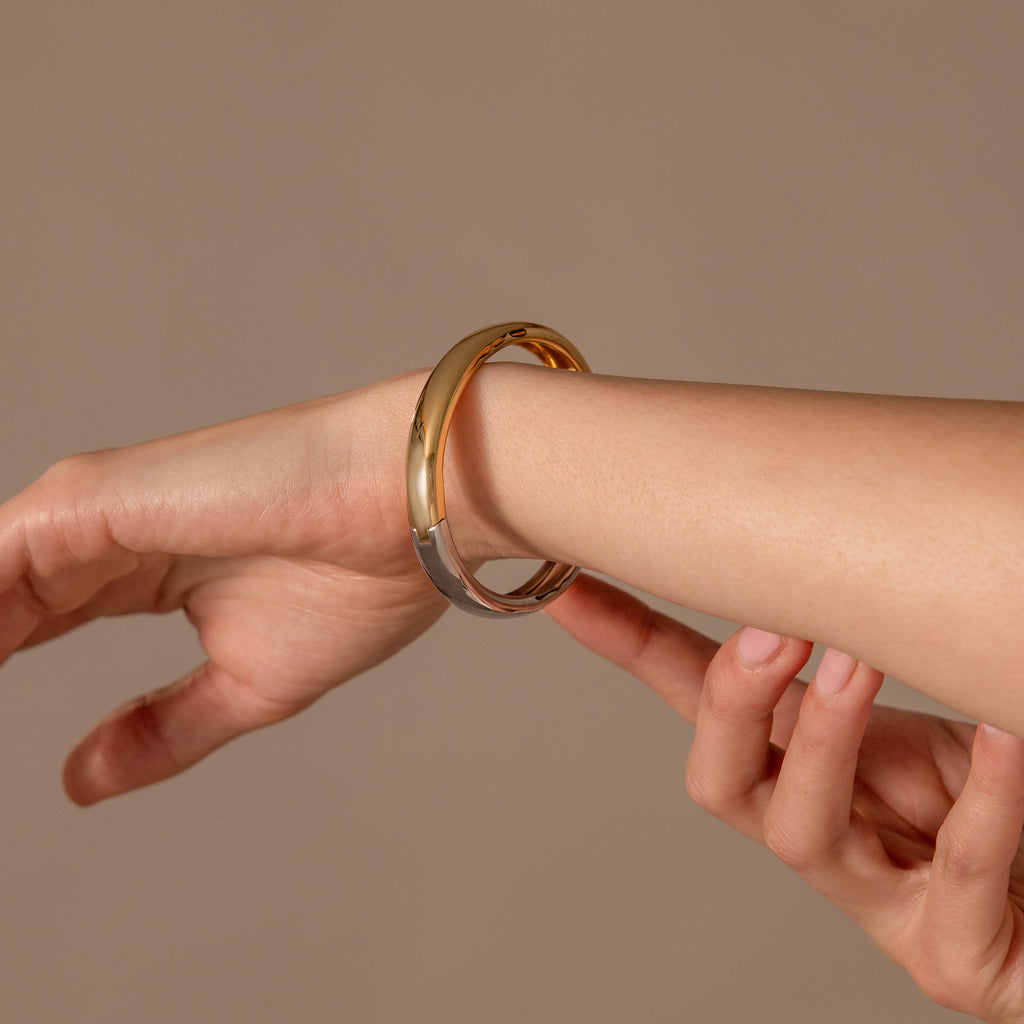 A hand slides the Mixed Metal Bangle—gold and silver intertwined—onto another wrist against a neutral background, highlighting its elegant minimalist design.