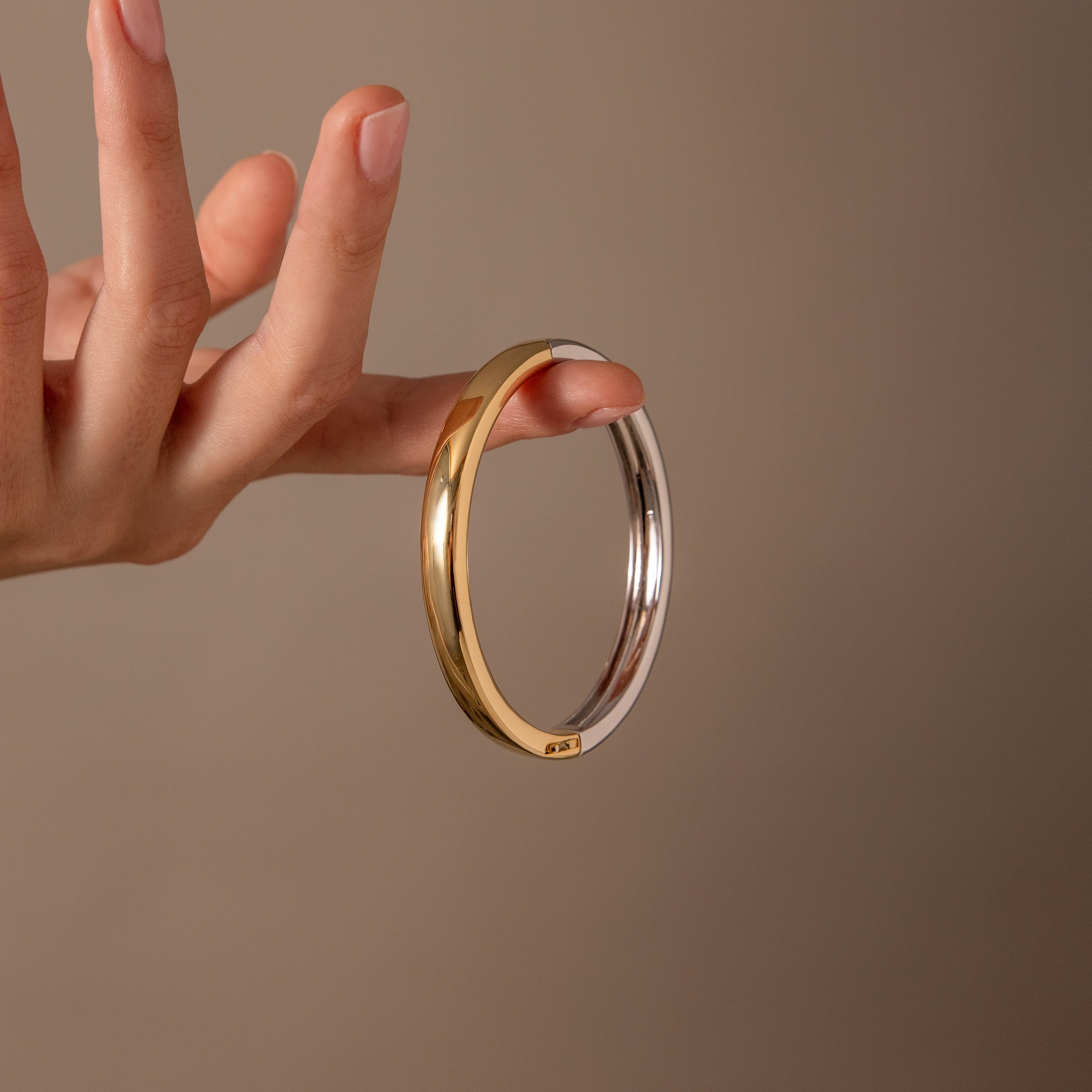 A hand displays the Mixed Metal Bangle—a minimalist gold and silver bracelet—set against a simple beige background.
