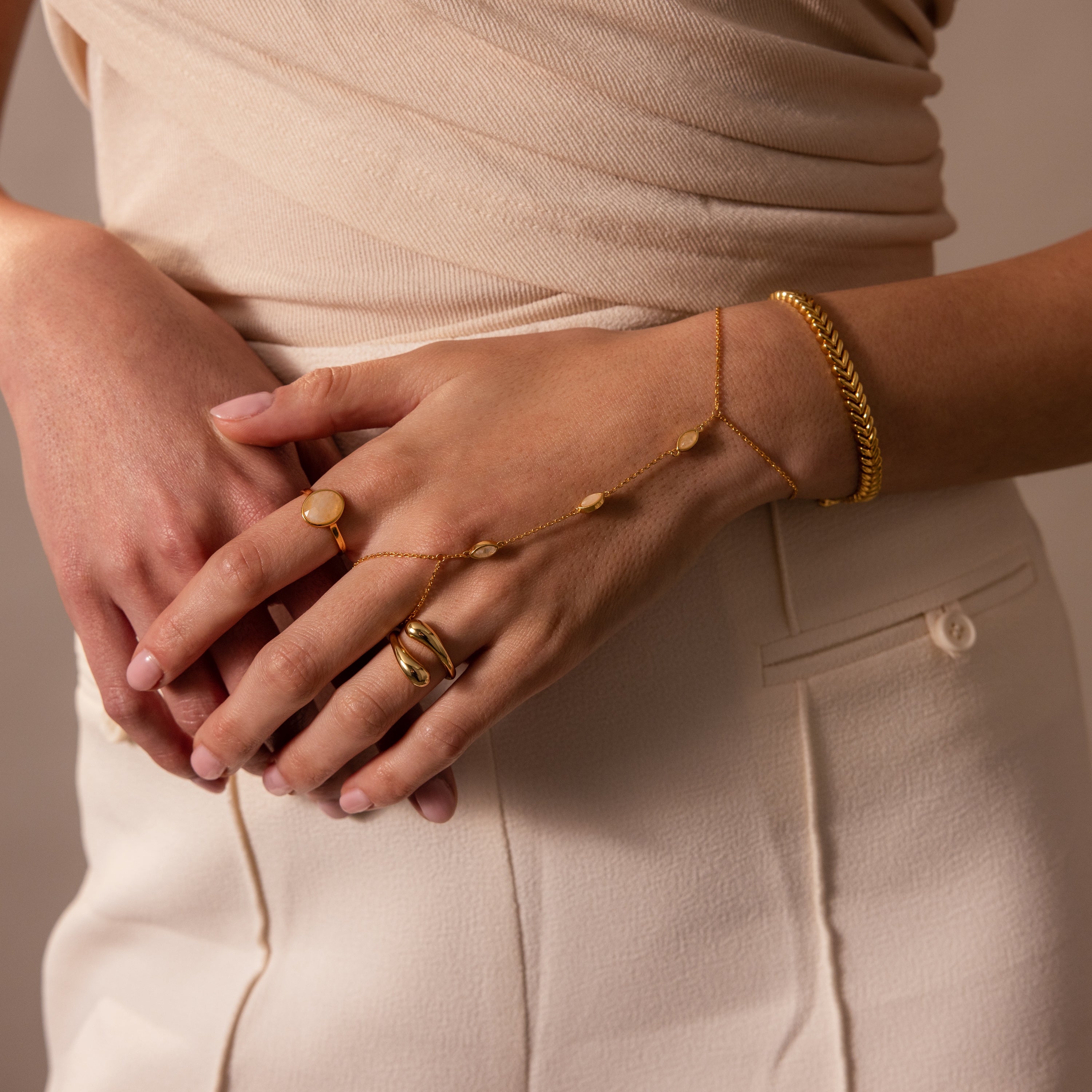 A woman wears the Moonstone Hand Chain Bracelet in 18K Gold over a cream top and skirt, her hands gently crossed.