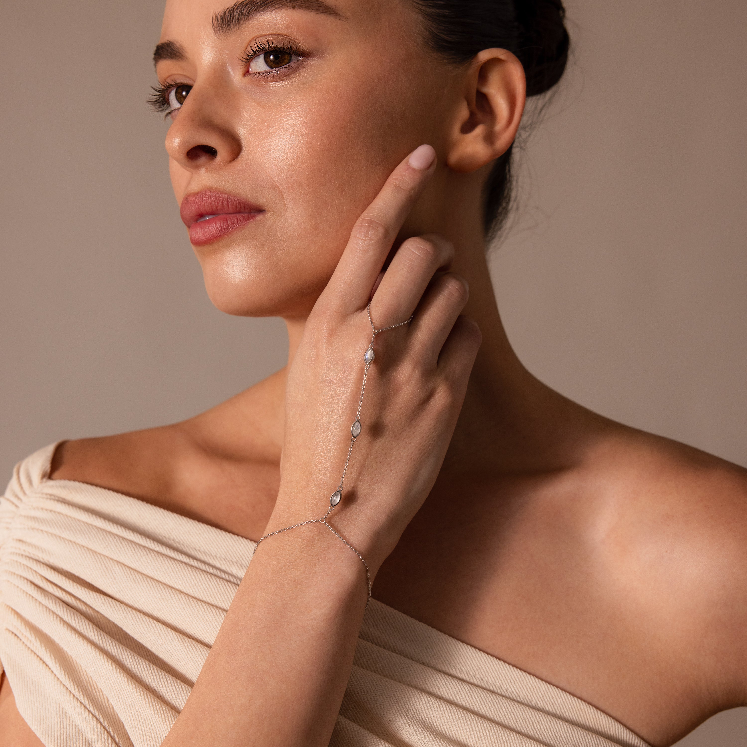 A woman in an off-shoulder top gazes thoughtfully to the side, showcasing the Moonstone Hand Chain Bracelet in Silver on her wrist.