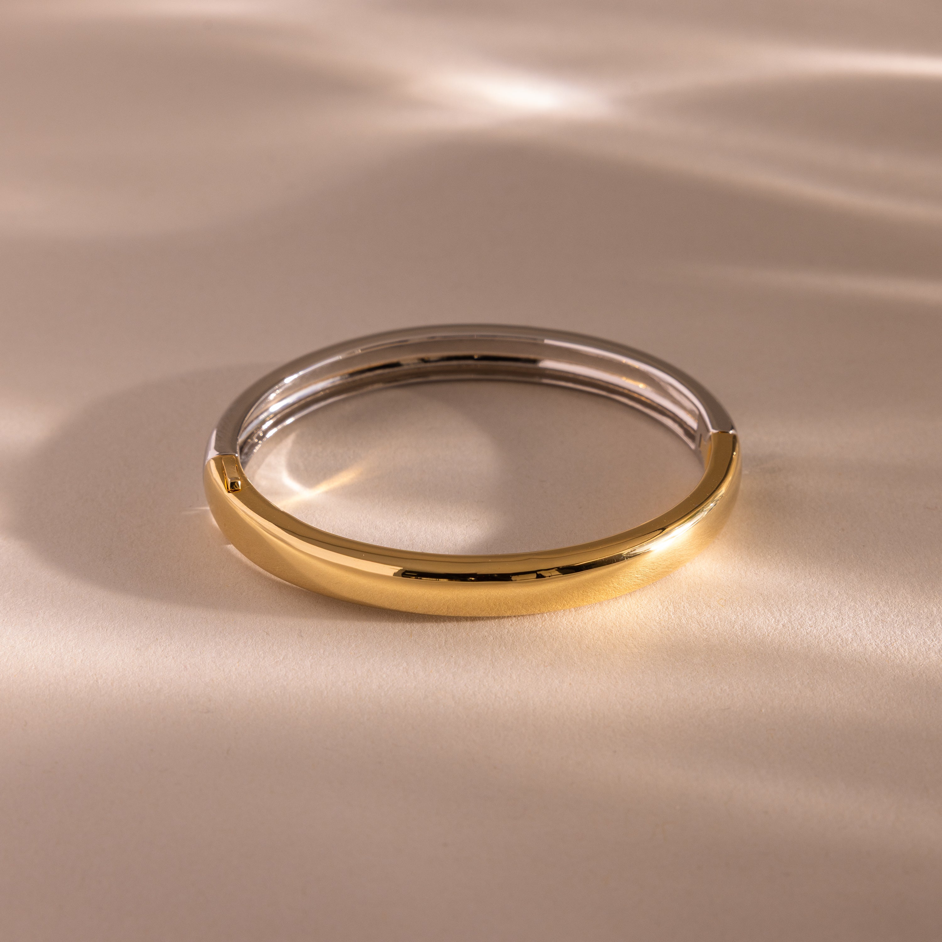 The Mixed Metal Bangle is a minimalist jewelry piece featuring silver and gold halves resting on a light, softly textured surface.