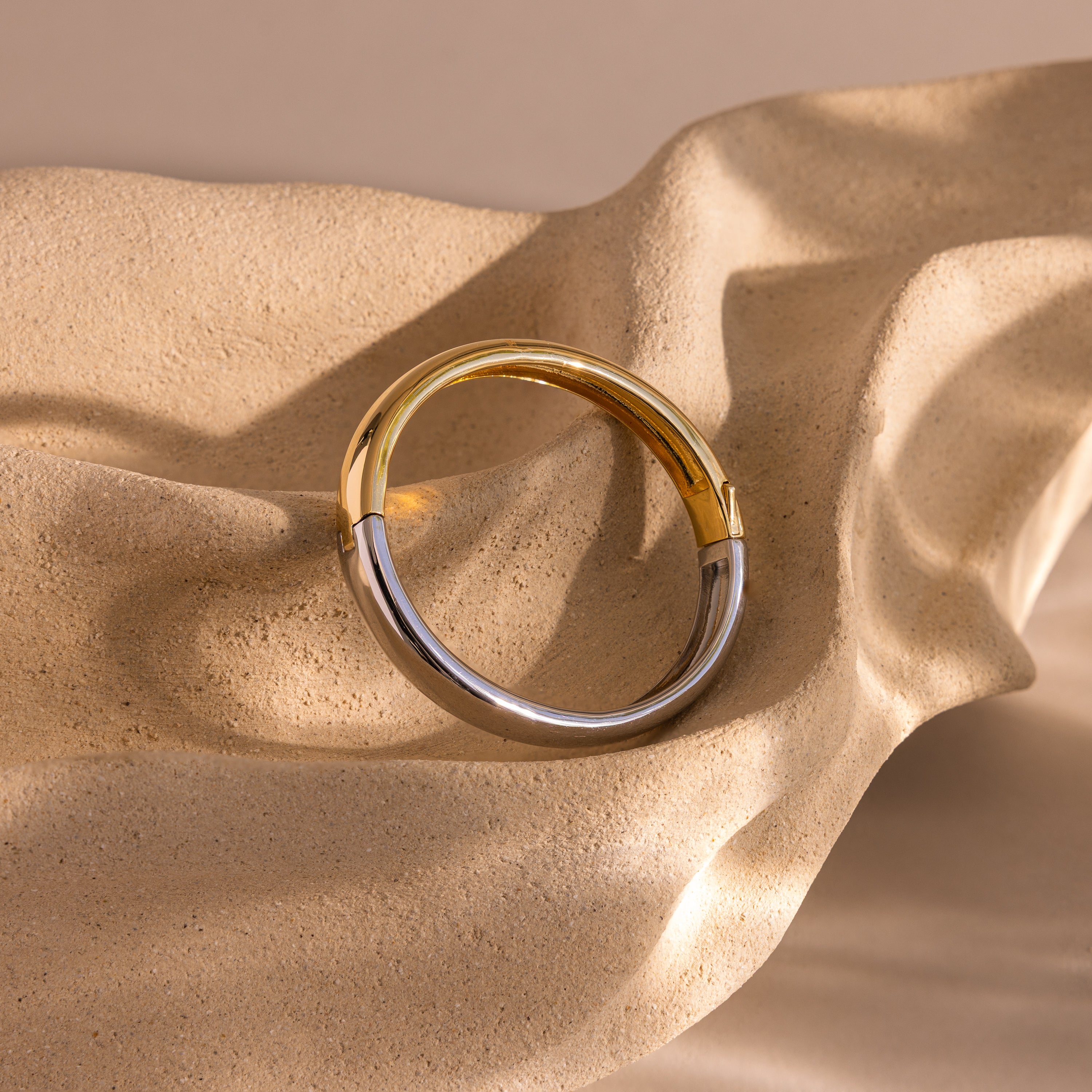 The Mixed Metal Bangle, a minimalist jewelry piece in gold and silver tones, is elegantly displayed on a textured beige wavy surface.