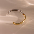 The Mixed Metal Bangle is an open cuff bracelet with half gold and half silver tones on a glossy beige surface, offering soft reflections—ideal for fans of minimalist jewelry.