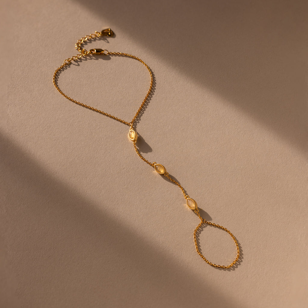 The Moonstone Hand Chain Bracelet in 18K Gold features three oval moonstone accents, shown on a beige background with soft lighting.