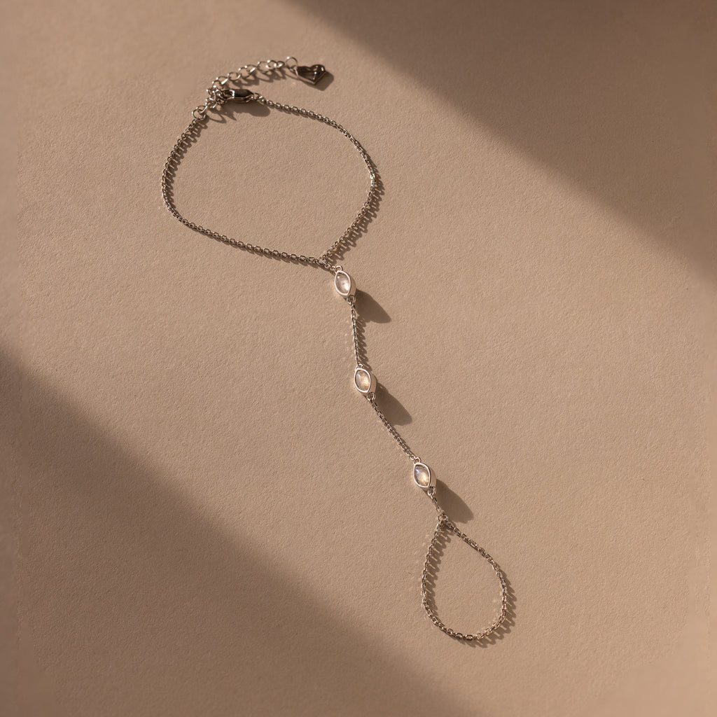 The Moonstone Hand Chain Bracelet in Silver features three oval gemstones and is shown on a beige background with gentle shadows.