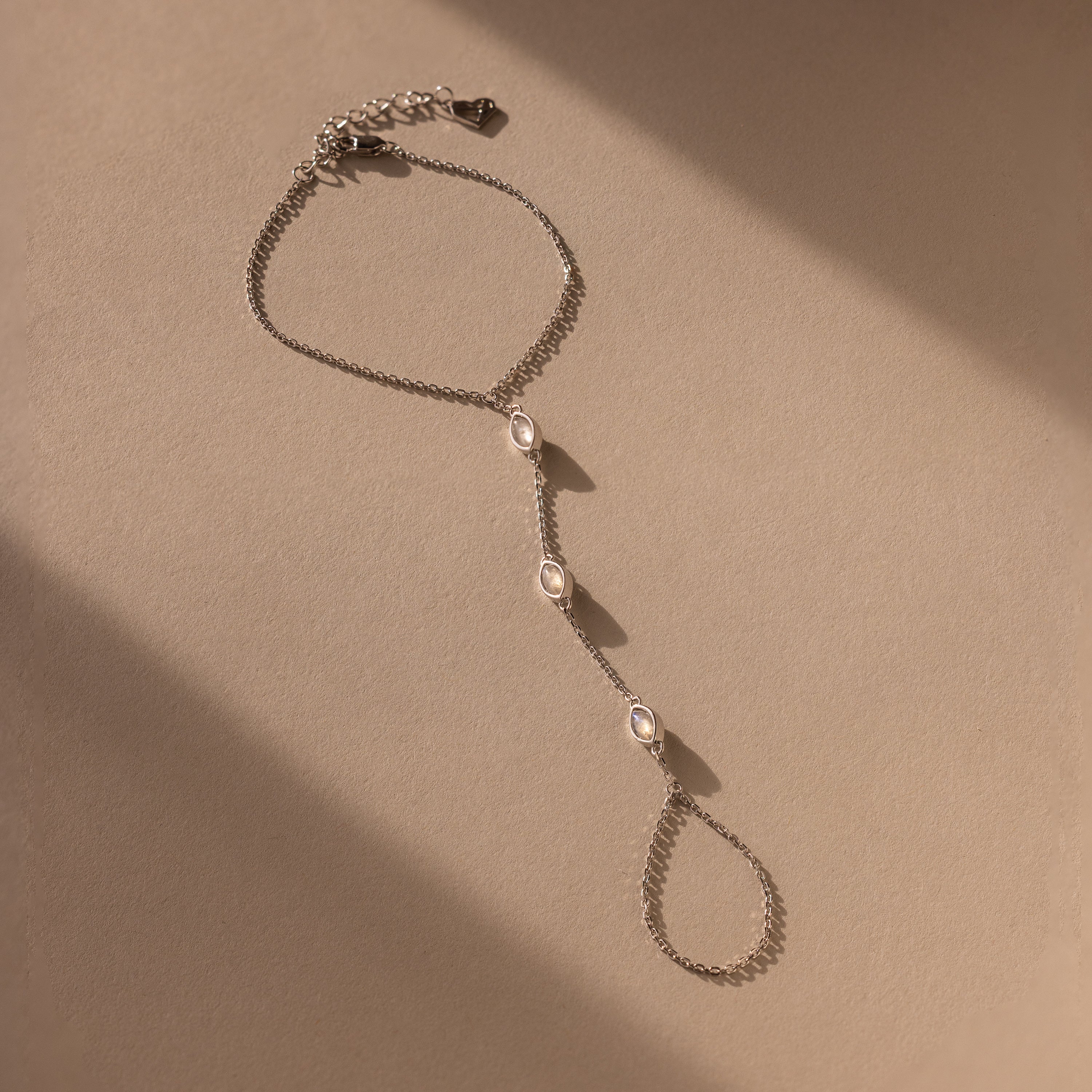 The Moonstone Hand Chain Bracelet in Silver features three oval gemstones and is shown on a beige background with gentle shadows.