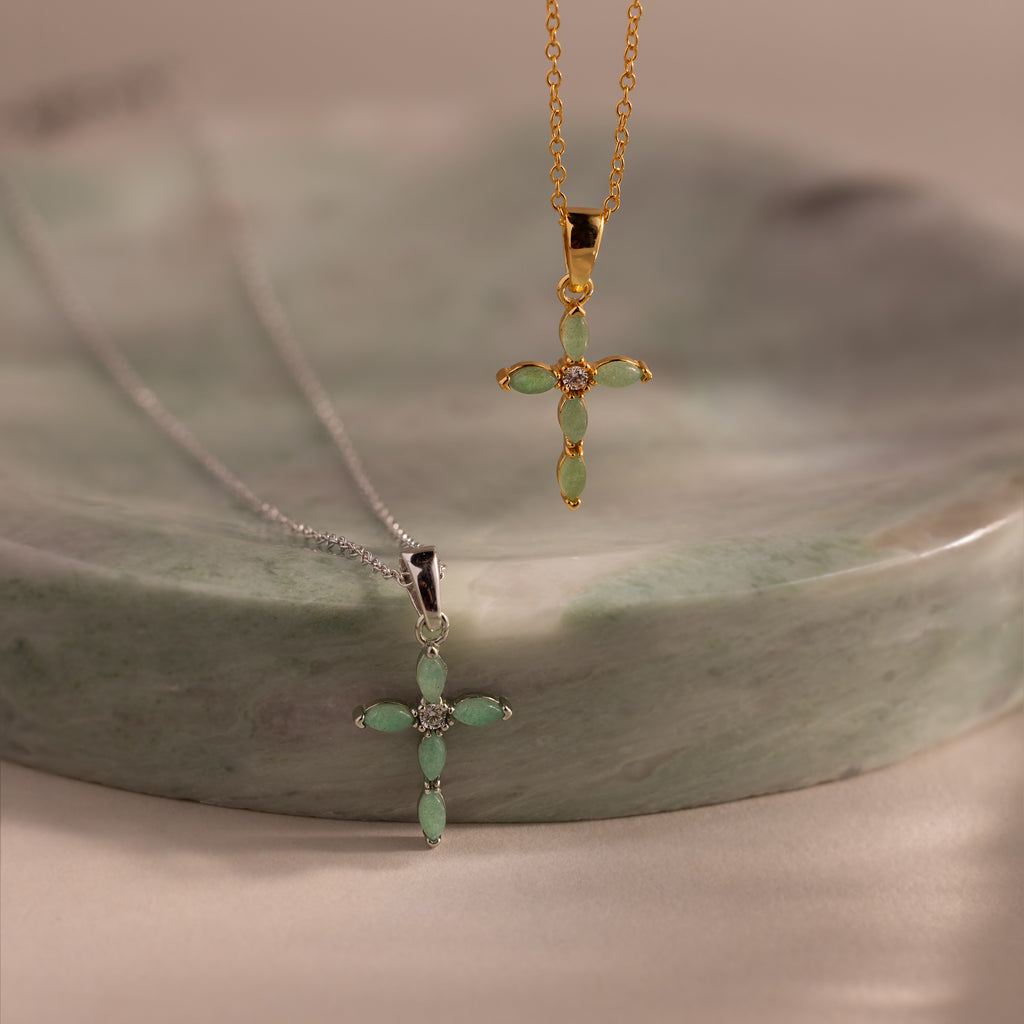 Close-up of two jade cross pendant necklaces in gold and silver, each featuring marquise-cut green stones and a CZ diamond center, displayed on a green stone dish.