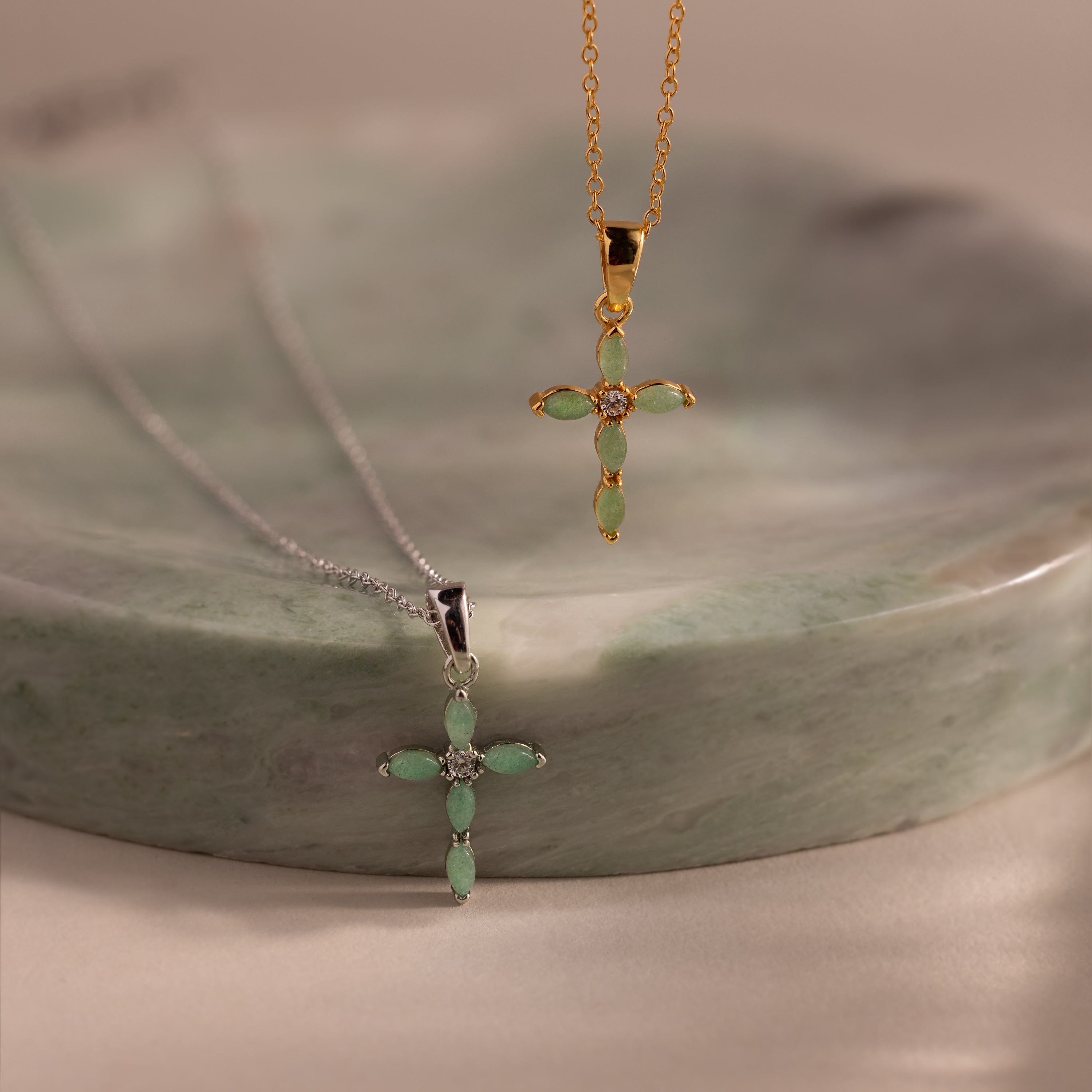 Close-up of two jade cross pendant necklaces in gold and silver, each featuring marquise-cut green stones and a CZ diamond center, displayed on a green stone dish.