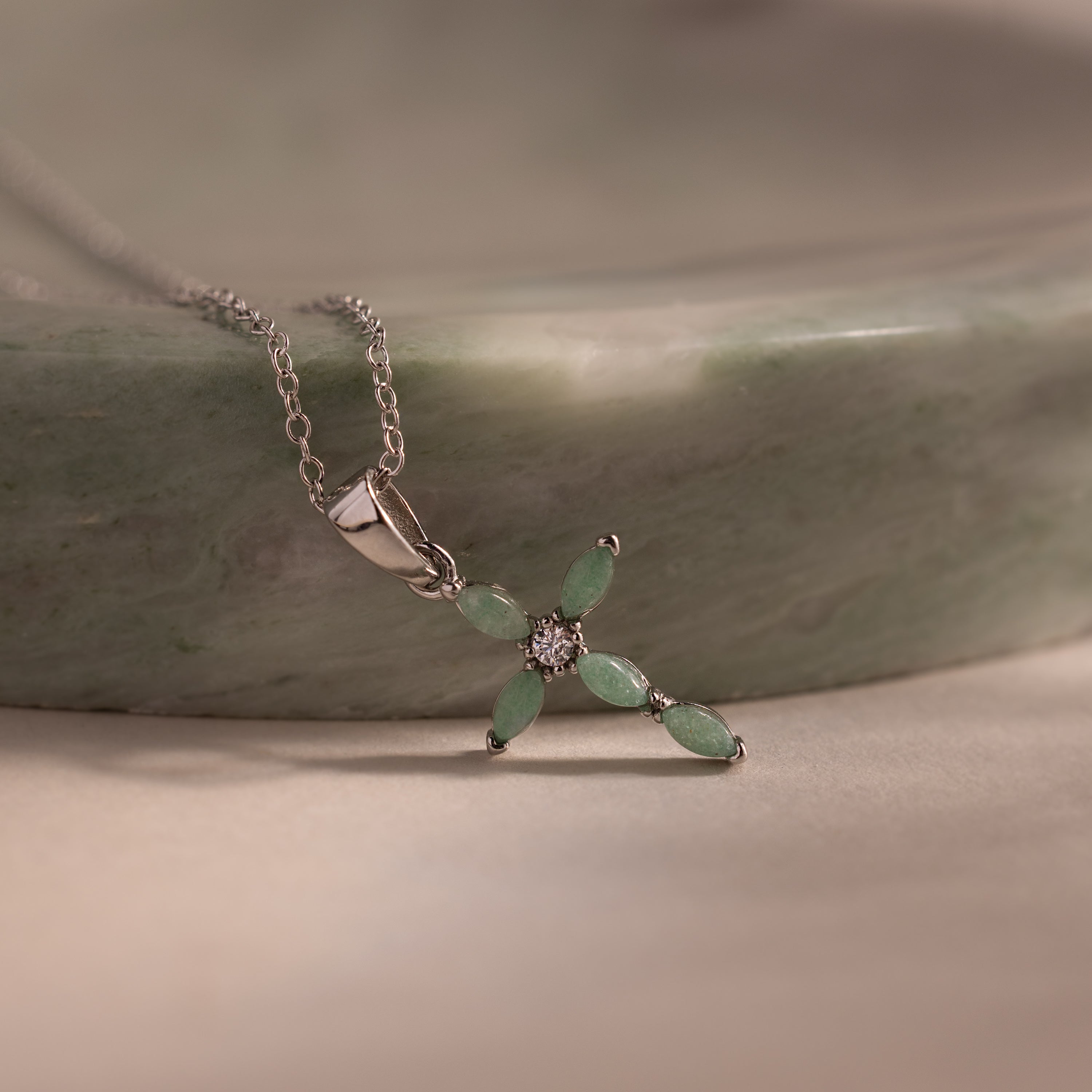 Silver chain necklace featuring a marquise jade stone cross pendant with a sparkling CZ diamond, displayed on a soft green stone surface.