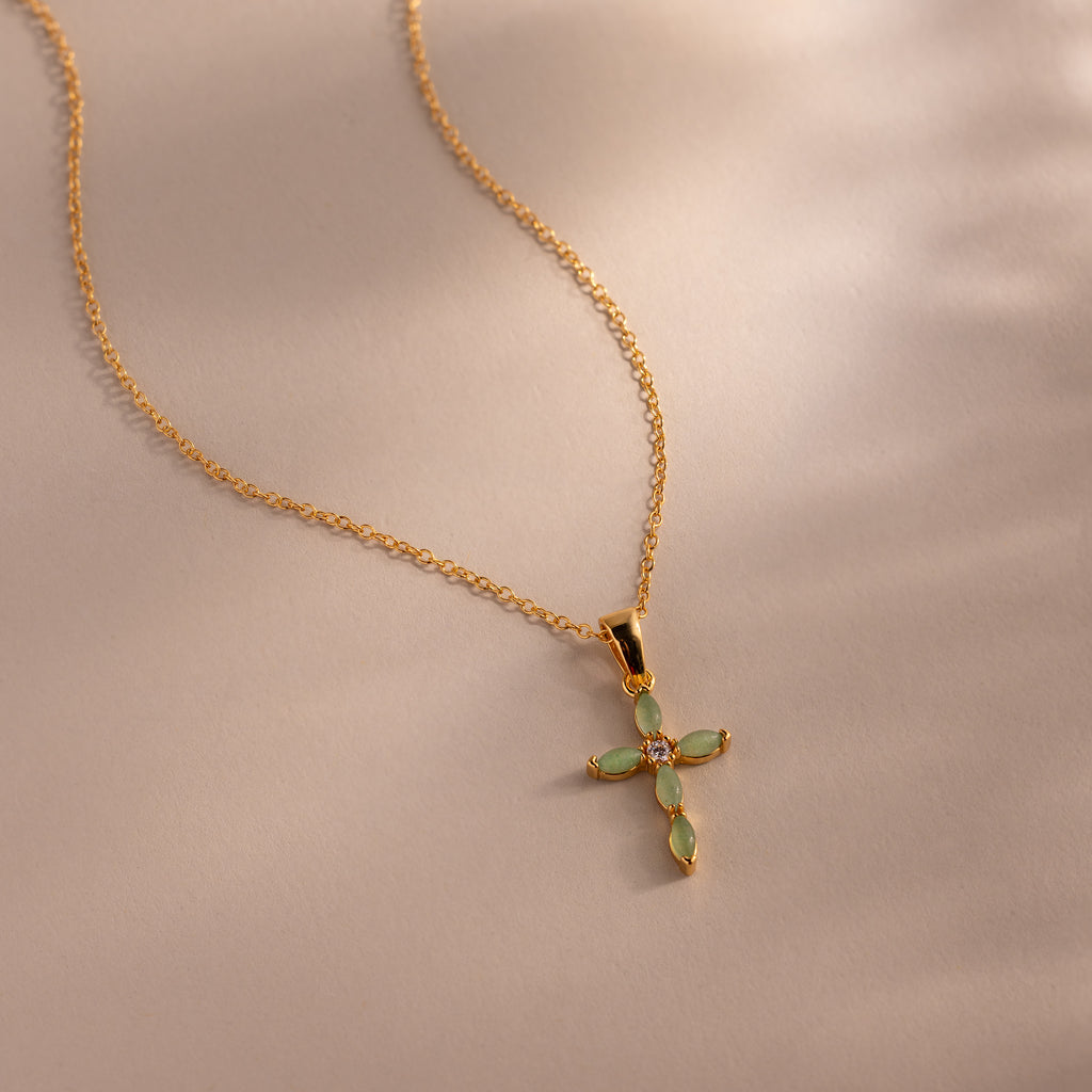 Flat-lay of a gold necklace with a jade cross pendant, featuring marquise-cut green stones and a CZ diamond center resting on a neutral background.