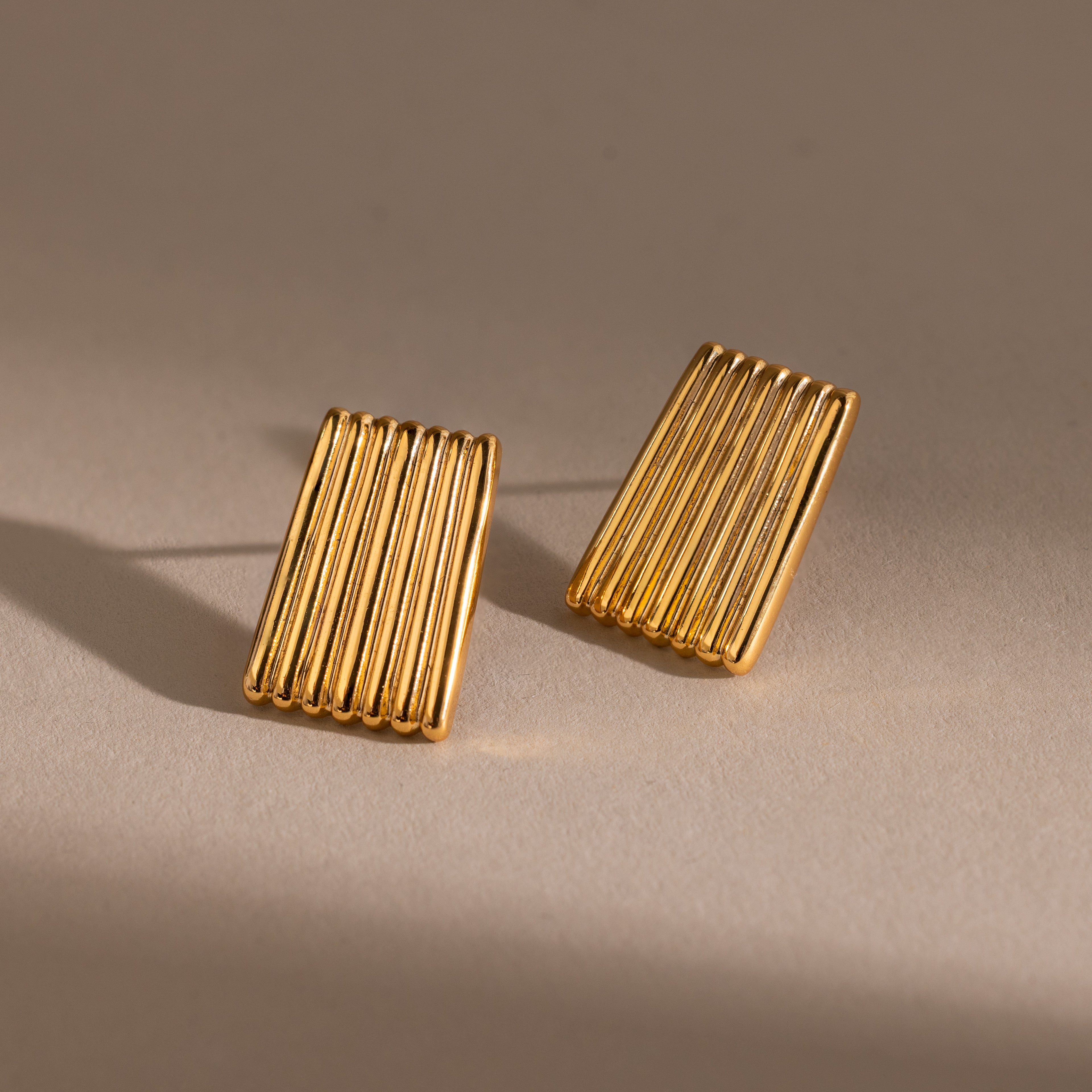 Ribbed Earrings—rectangular gold earrings with parallel ridges, displayed on a beige surface with soft shadows—make a bold statement in contemporary jewelry.