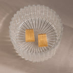 Ribbed Earrings in 18K Gold with a textured column design rest in a clear glass dish on a beige surface, adding a touch of contemporary jewelry style.