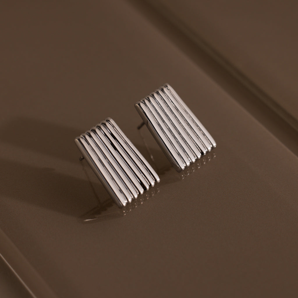 Ribbed Earrings in White Gold featuring a textured column design with vertical ridges, displayed on a brown textured surface—an elegant piece of contemporary jewelry.