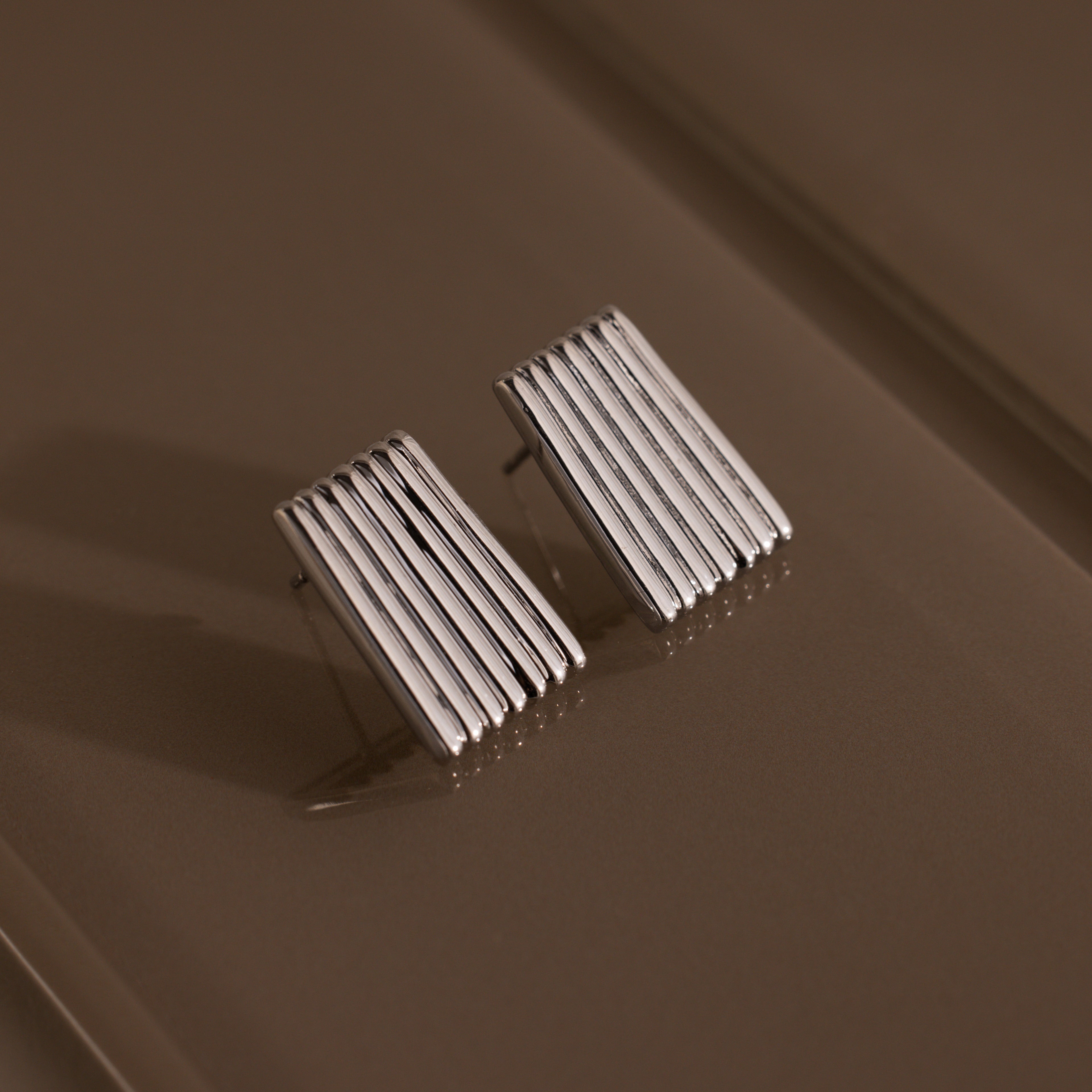 Ribbed Earrings in White Gold featuring a textured column design with vertical ridges, displayed on a brown textured surface—an elegant piece of contemporary jewelry.
