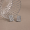Two rectangular Ribbed Earrings in White Gold, featuring a textured column design, are displayed next to a clear ridged glass object on a beige surface.