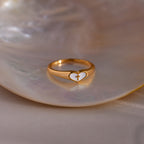 Gold heart-shaped ring with a white enamel face and small gold cross, displayed on a seashell dish.