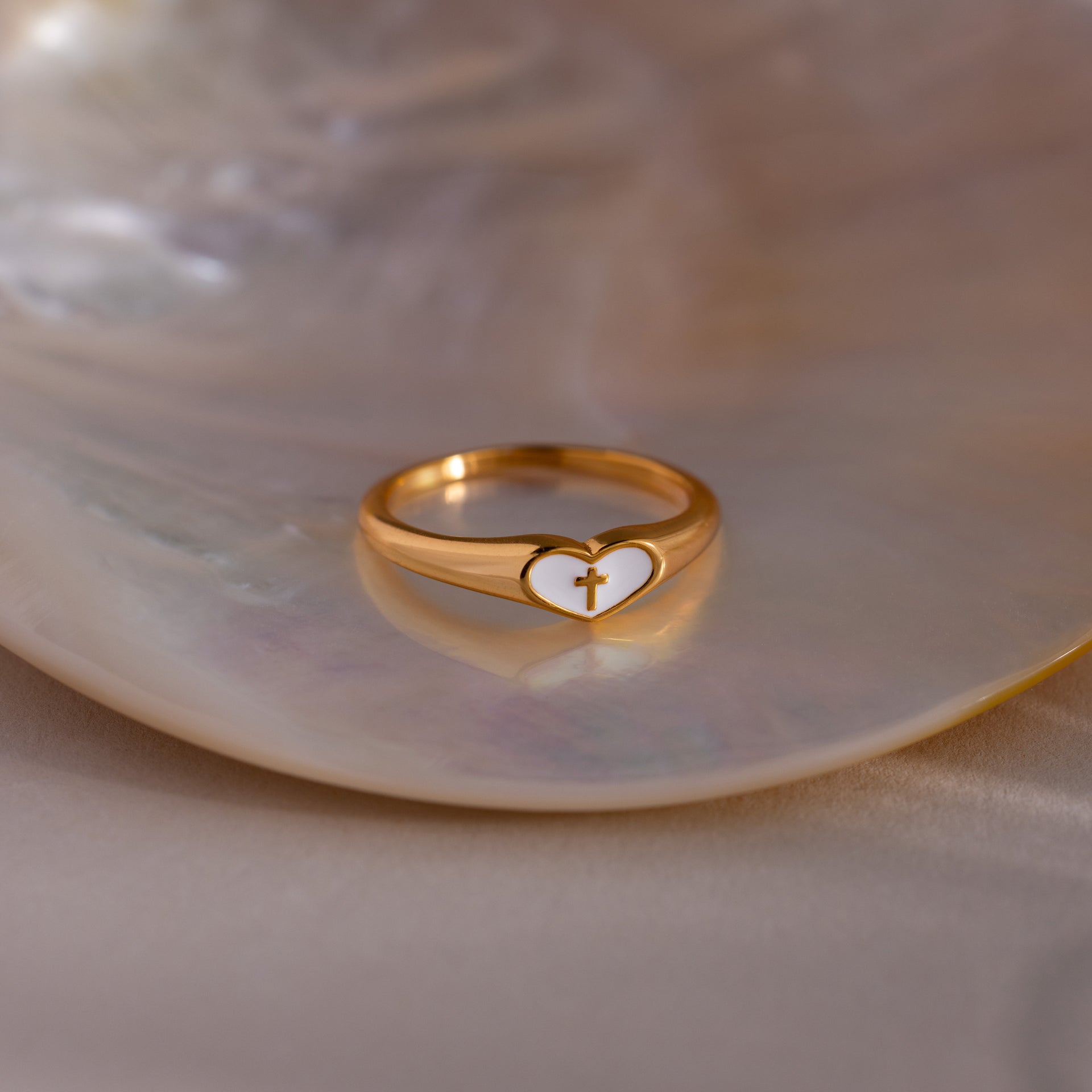Gold heart-shaped ring with a white enamel face and small gold cross, displayed on a seashell dish.