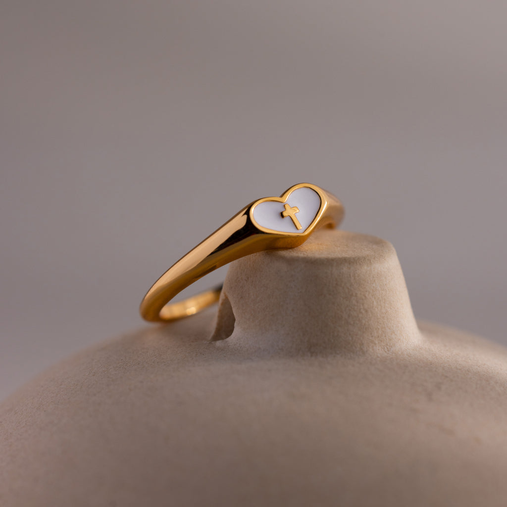 Close-up of a gold heart-shaped ring with a white enamel face and small gold cross.